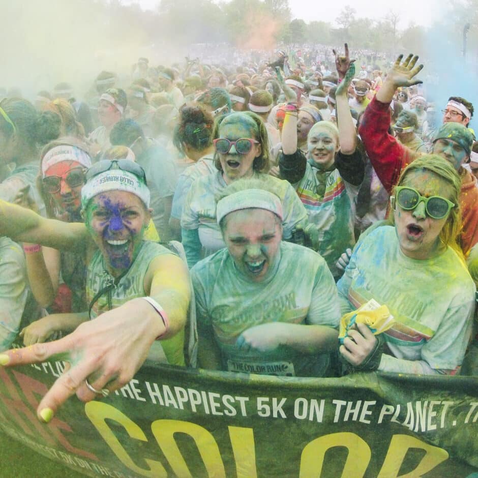 People wearing colorful powder and sunglasses at a vibrant outdoor color run event. They exude joy and excitement, surrounded by a festive atmosphere.