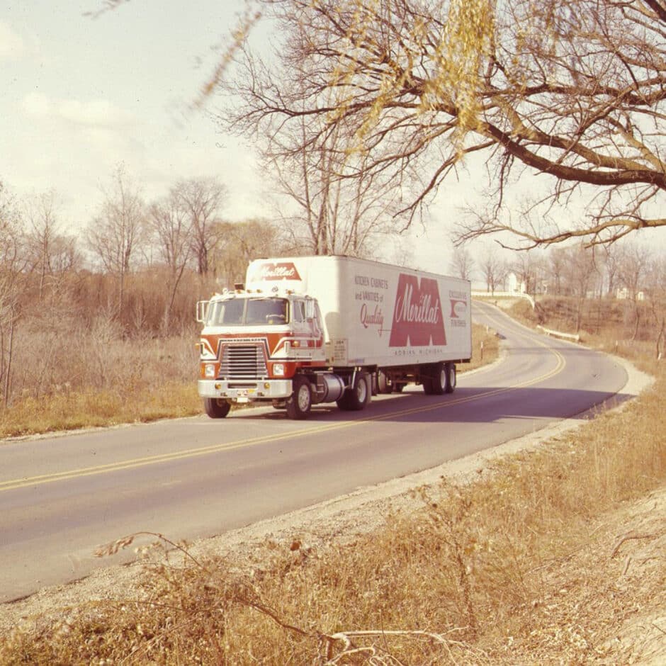 A vintage red and white truck with a "Moulin" logo drives along a curved rural road. Leafless trees line the path, evoking a calm, autumnal feel.