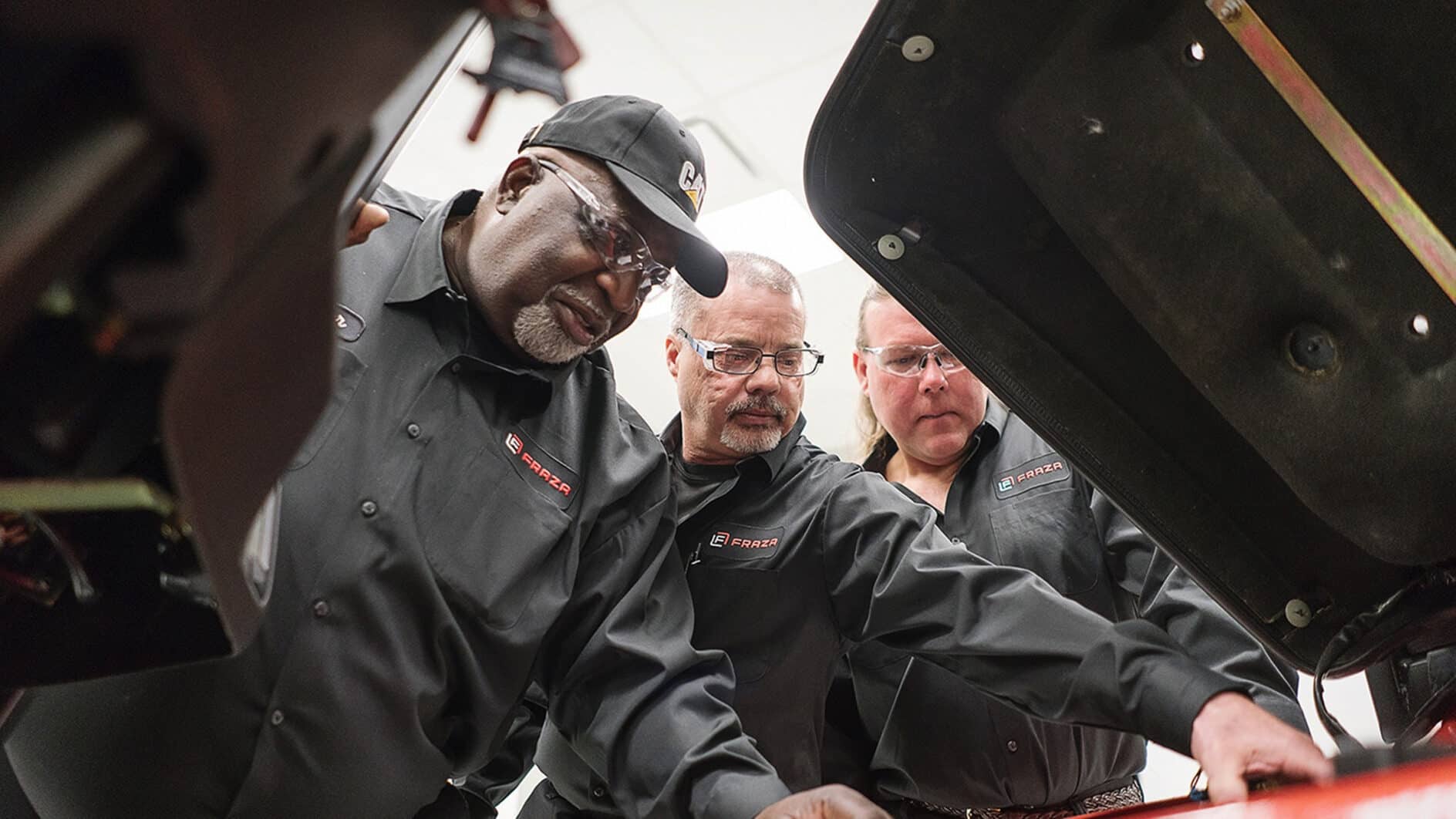 Three mechanics in black uniforms and safety glasses inspect a vehicle engine intently, conveying focus and teamwork in a garage setting.