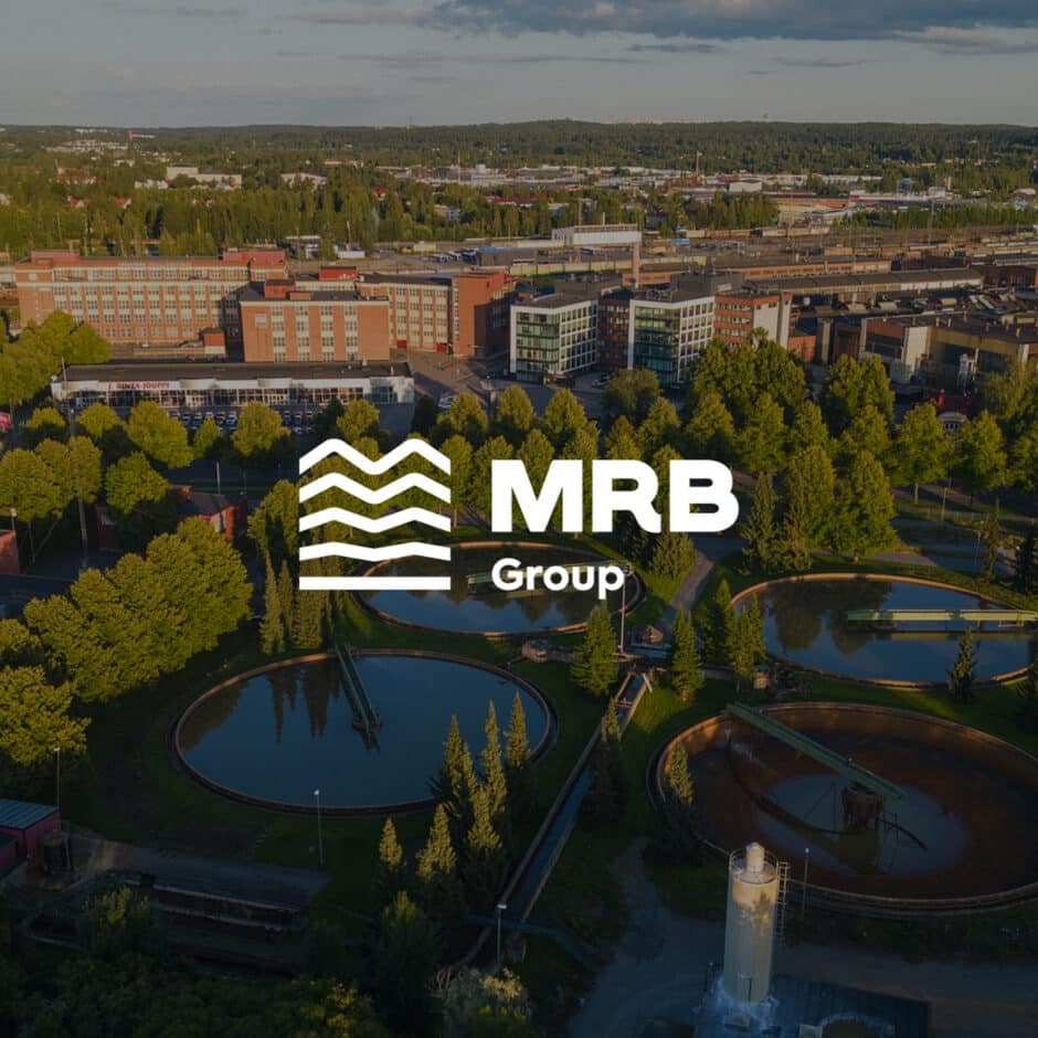 Aerial view of a water treatment facility with circular tanks surrounded by trees and buildings. The MRB Group logo is prominently displayed in the center.
