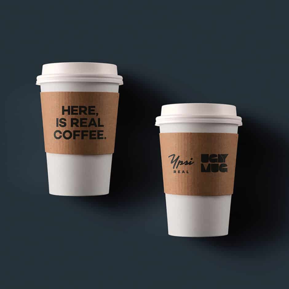 Two white coffee cups with brown sleeves on a dark gray background. The left cup reads "Here, is real coffee." The right cup displays two logos.