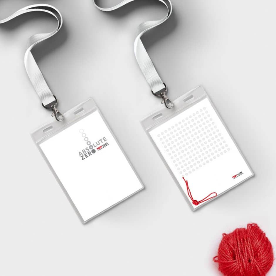 Two white lanyard cards, one with "Absolute Zero" and logos, the other with gray dots and a red string. A red yarn ball lies nearby on a white backdrop.