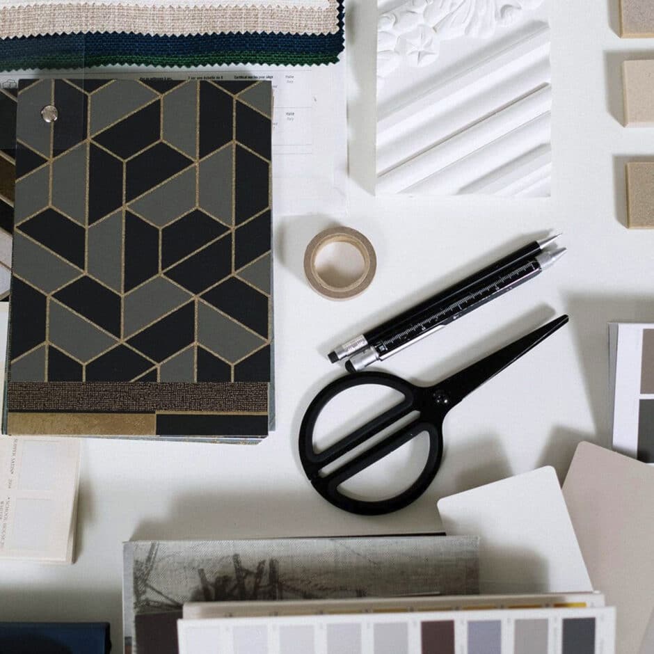 Aerial view of a design workspace with black scissors, geometric-patterned booklet, pens, fabric samples, tape, and texture swatches on a white surface.