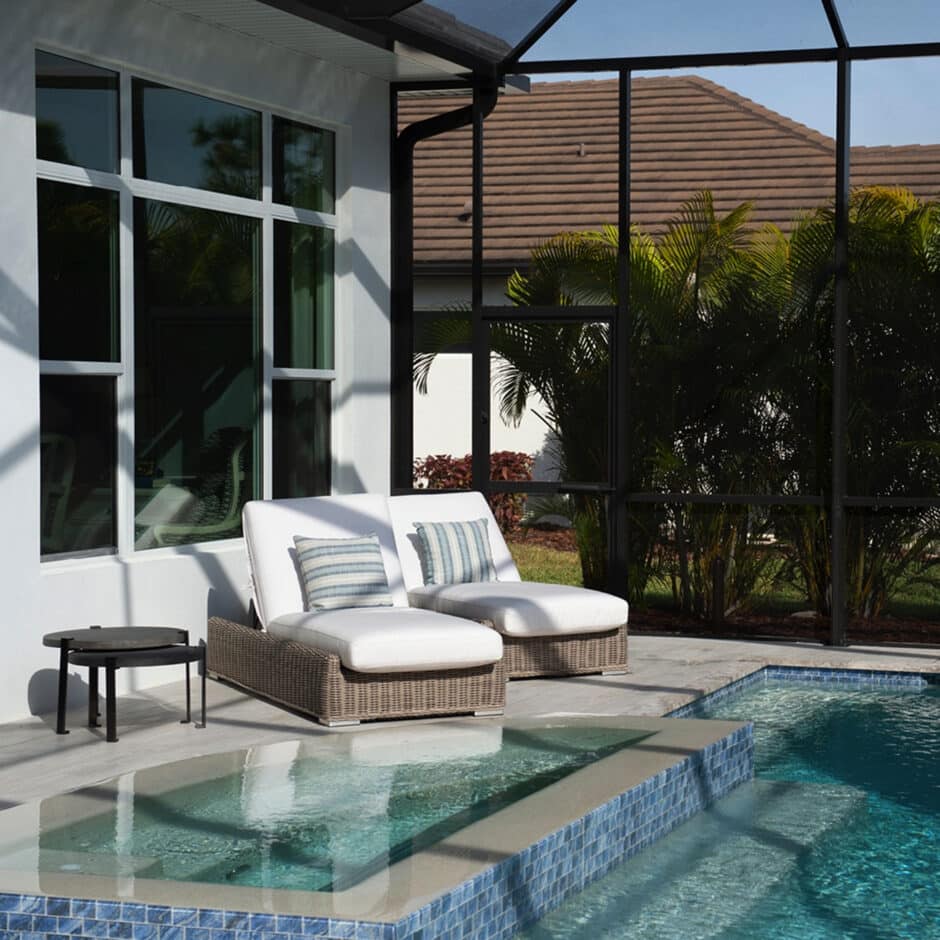 A sunlit patio with two cushioned wicker loungers, a small side table, and clear blue pool. Palm trees and a screened enclosure complete the serene setting.