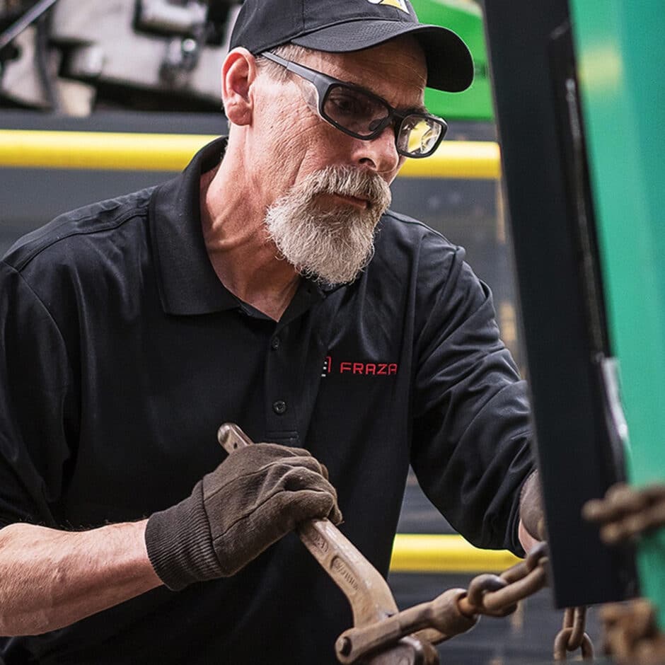 A man with a beard and glasses, wearing a black cap and shirt, uses a wrench on machinery. He is focused and appears to be in an industrial setting.