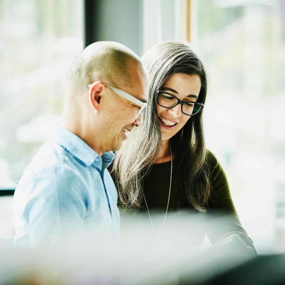 Two people smiling at something off-camera, with soft natural light coming through the window. The atmosphere is friendly and collaborative.