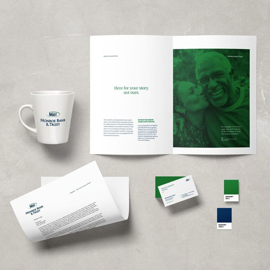 Open brochure with a smiling couple and text, "Here for your story, not ours." Nearby are branded items: a Monroe Bank & Trust mug, letterhead, business cards, and color swatches in green and blue.