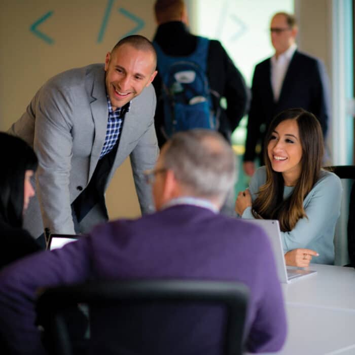 A group of professionals gather around a table in an office setting. A man in a gray suit leans forward, smiling warmly, while a woman sits, smiling back. Laptops are open, indicating a collaborative meeting. The atmosphere is friendly and engaged.