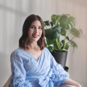 A woman with dark hair and a bright smile sits in front of a leafy plant. She's wearing a blue gingham blouse, creating a cheerful and relaxed tone.
