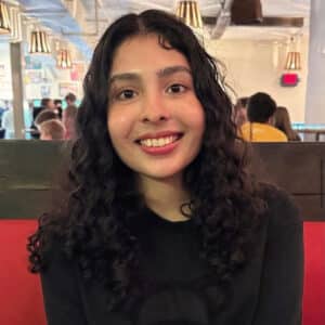 Young woman with curly hair smiling warmly, seated in a casual dining setting with soft lighting. Background shows people and hanging lamps.