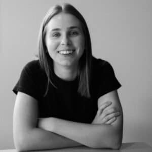 Smiling woman with straight hair, arms crossed on a table, wearing a black T-shirt. The black and white photo conveys a friendly, relaxed tone.