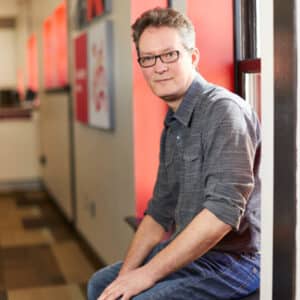 A man with glasses and a gray shirt sits casually by a window with red accents. The space exudes a modern, relaxed atmosphere.