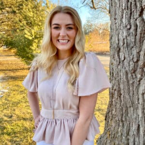 Smiling woman with long blonde hair in a light blouse stands near a tree. Background shows a sunny park with green and bare trees.