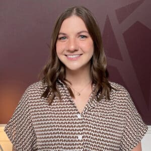 Young woman with brown hair smiles against a maroon background with geometric patterns. She wears a patterned blouse. The tone is warm and welcoming.