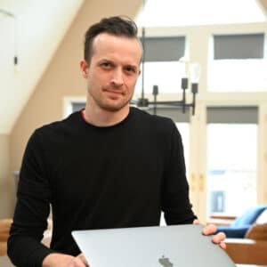 A person in a black sweater holds a laptop, standing in a modern room with large windows and minimalist decor, conveying a professional tone.