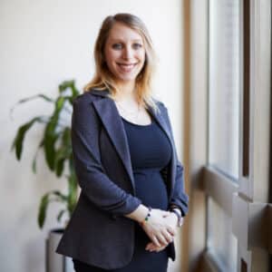 A smiling woman stands by a large window, wearing a black dress and gray blazer. A plant is in the background, suggesting a professional setting.