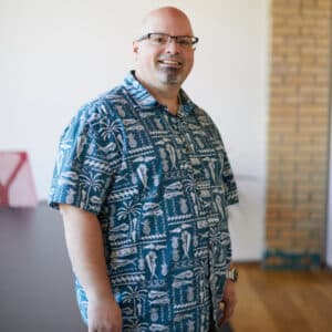 Smiling man wearing a blue patterned shirt stands in a modern room with brick walls. Warm, relaxed atmosphere with natural light.