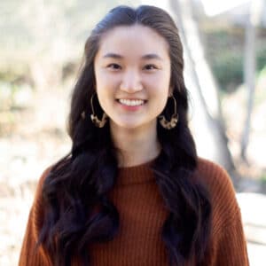 Smiling woman with long dark hair and hoop earrings, wearing a brown sweater. She is outdoors with a blurred natural background, conveying warmth.