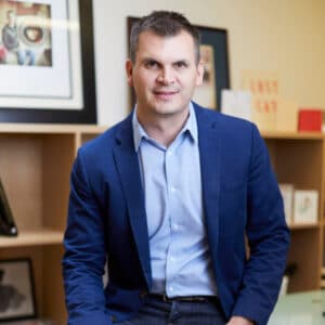 Man in a blue blazer and light blue shirt sits on desk edge, smiling. Background features wooden shelves with books and framed pictures, conveying a professional yet relaxed atmosphere.