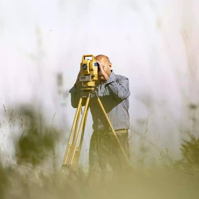 A surveyor in a checkered shirt uses a yellow theodolite on a tripod in a grassy field. The scene conveys precision and focus amid soft natural surroundings.
