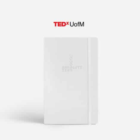 White notebook embossed with "Absolute Zero" and circles, on a clean surface. Overhead text reads "TEDxUofM" in a bold, modern font. Minimalist and professional tone.