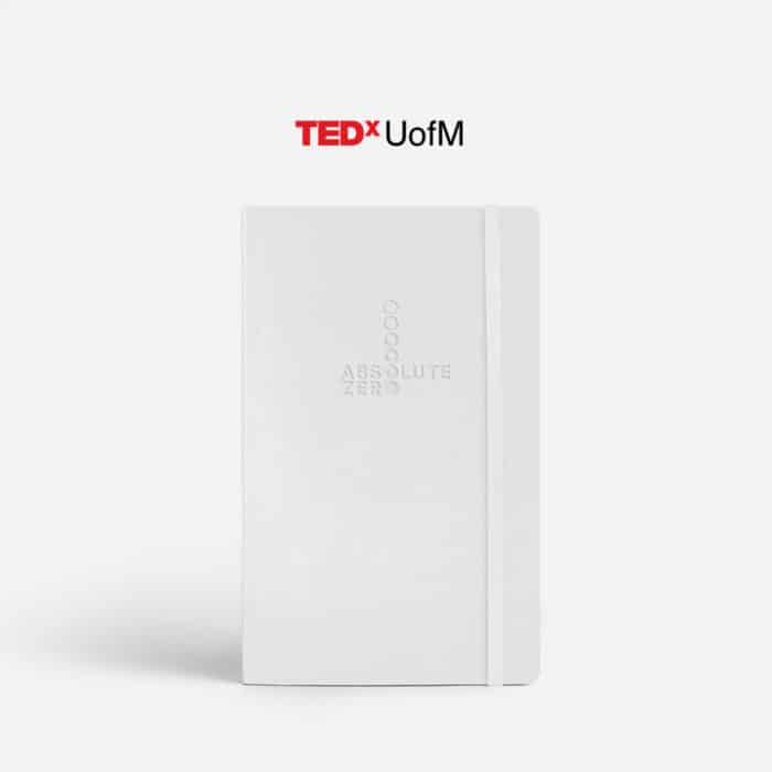 White notebook embossed with "Absolute Zero" and circles, on a clean surface. Overhead text reads "TEDxUofM" in a bold, modern font. Minimalist and professional tone.