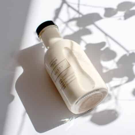 A bottle of nutty flavored drink lies on a white surface, casting a soft shadow. Delicate plant shadows add an artistic, serene touch.