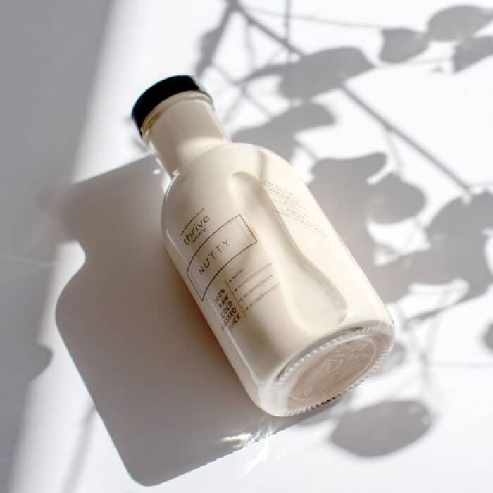 A bottle of nutty flavored drink lies on a white surface, casting a soft shadow. Delicate plant shadows add an artistic, serene touch.