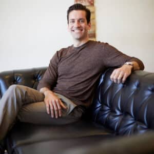 Smiling man seated on a black leather sofa, wearing a brown sweater and gray pants. The atmosphere appears relaxed and welcoming.