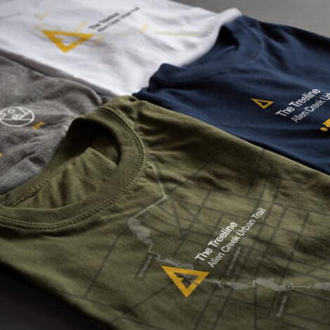 Three folded t-shirts in white, gray, and blue display abstract map designs and "The Treeline" text. The tone is modern and outdoorsy.