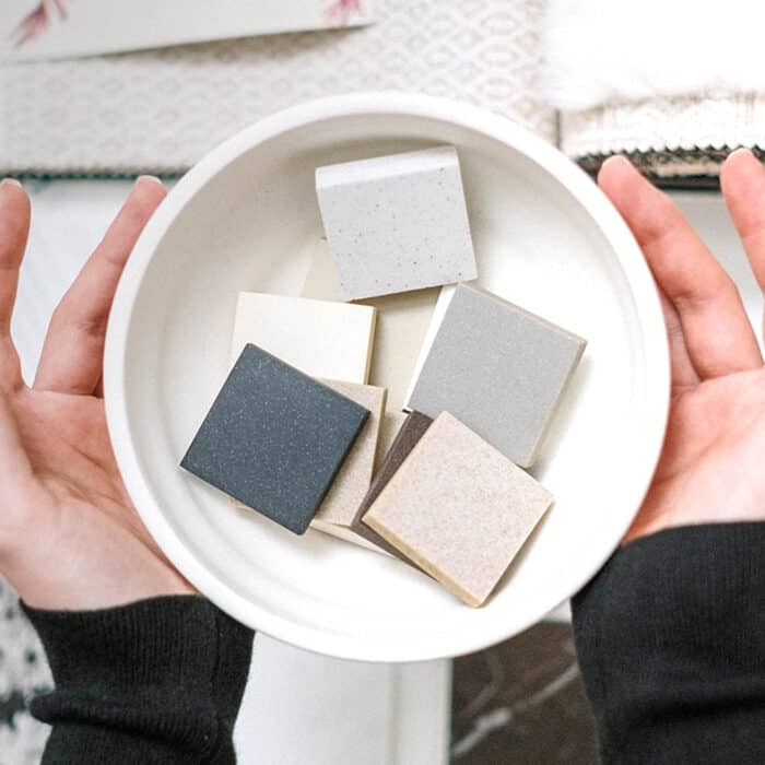 Two hands hold a white bowl containing six square tiles in neutral shades, like gray and beige, set against a soft, patterned background.
