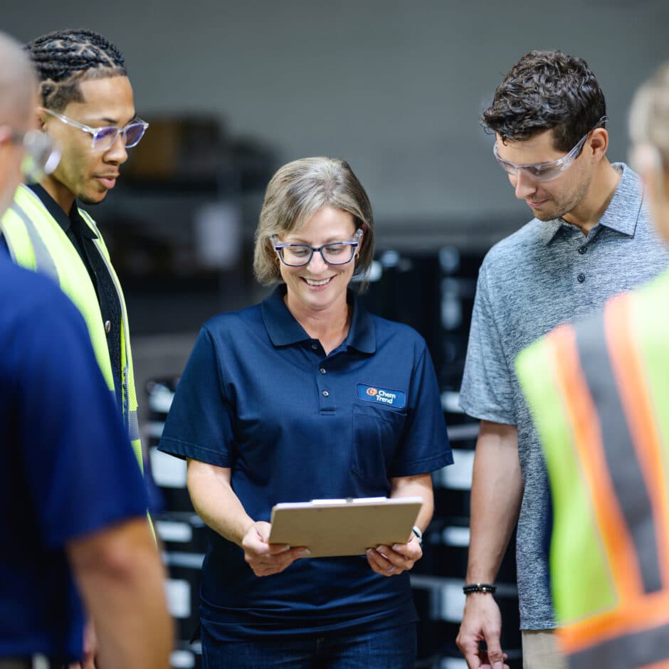 A woman in a blue polo with a logo reviews a clipboard, surrounded by three colleagues wearing safety glasses. They're in a workshop setting, appearing focused and collaborative.