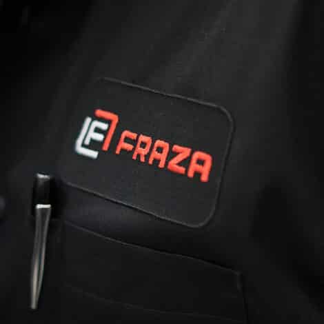 Close-up of a black shirt with a red and white embroidered "FRAZA" logo on a patch. A pen is clipped in the pocket, giving a professional tone.