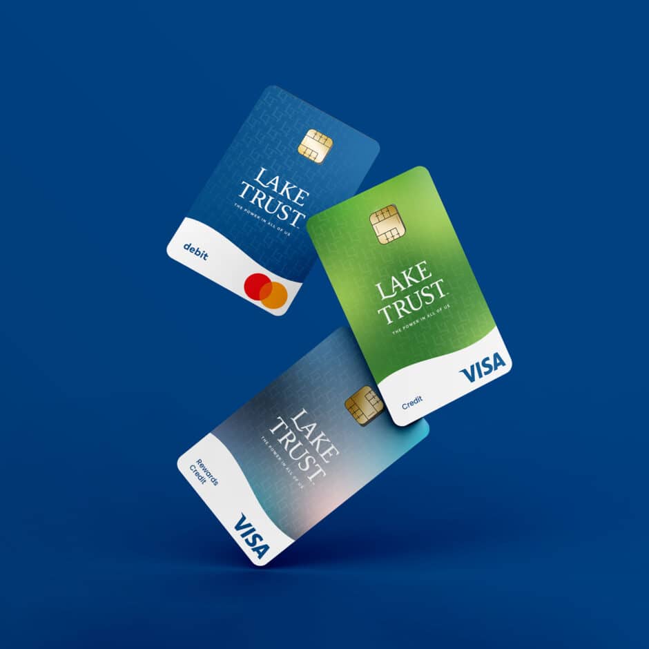 Three credit cards labeled "Lake Trust" are floating against a blue background. The cards are blue for Mastercard debit, green for Visa credit, and a gradient for Visa rewards.