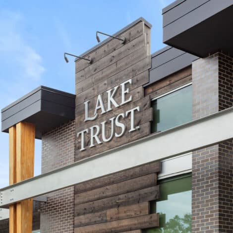 Modern building facade with "Lake Trust" sign on rustic wooden paneling, accented by glass windows and brickwork against a clear blue sky.