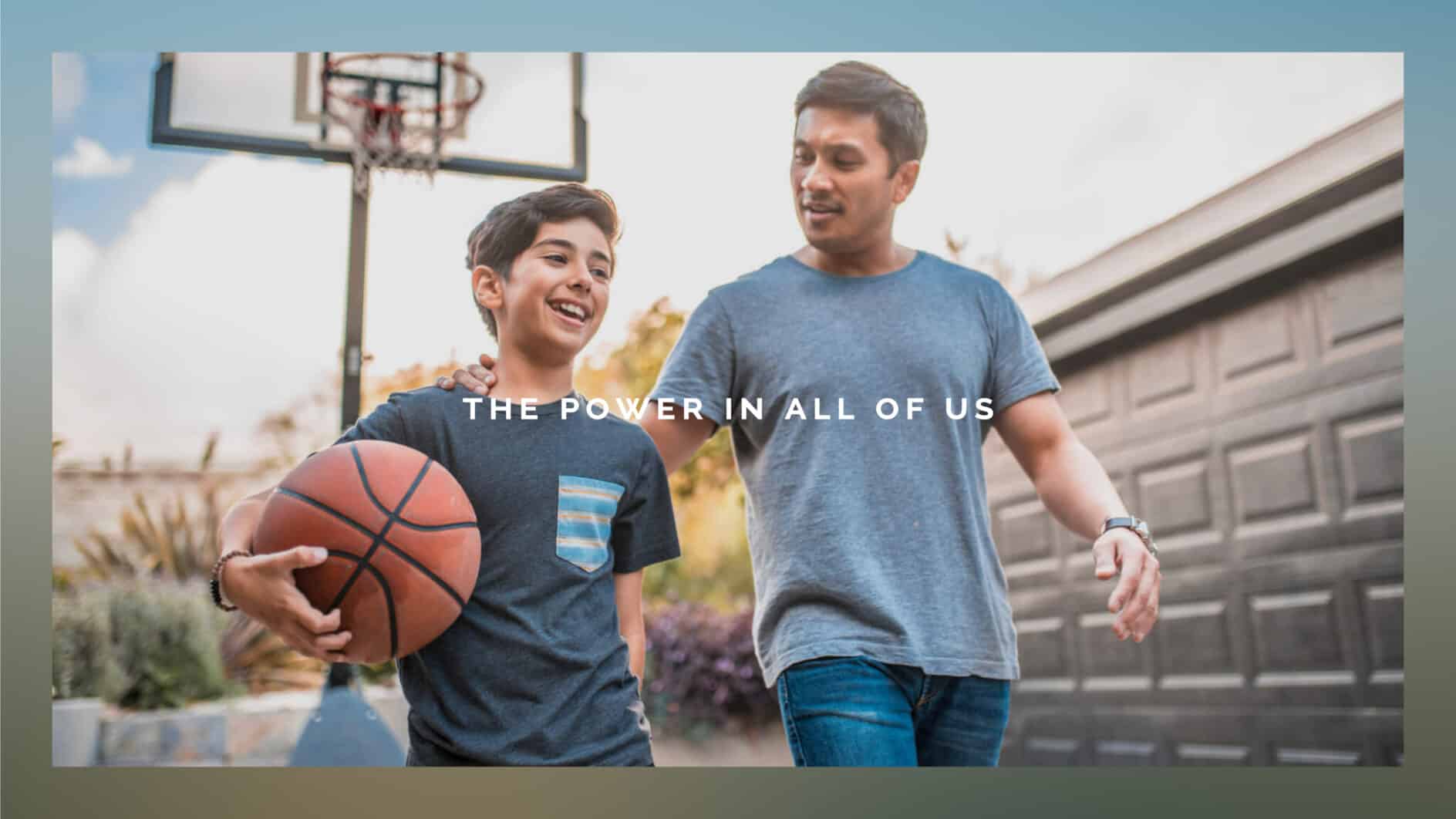 A father and son share a joyful moment outdoors. The son holds a basketball, and both are smiling. Text overlay reads, "The Power in All of Us."