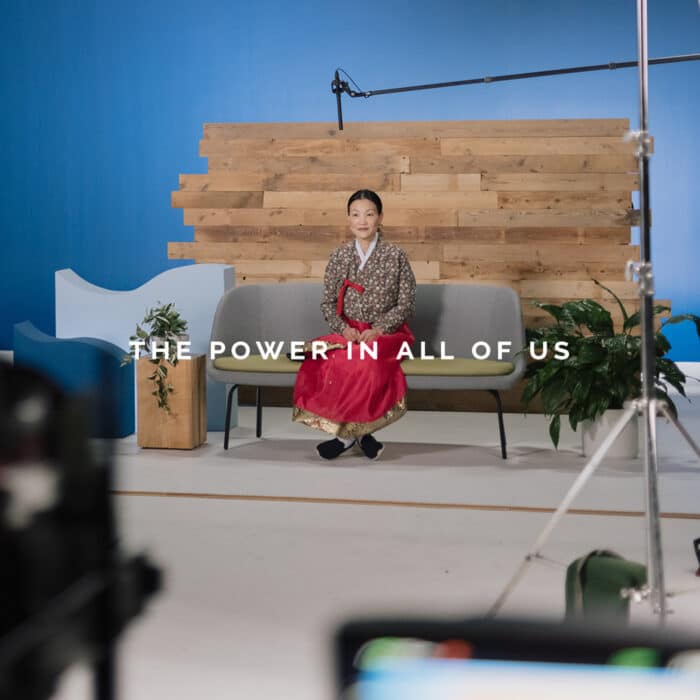 A person in traditional Korean attire sits on a gray sofa against a wooden backdrop. Text reads "The Power in All of Us." The setting is calm and professional.