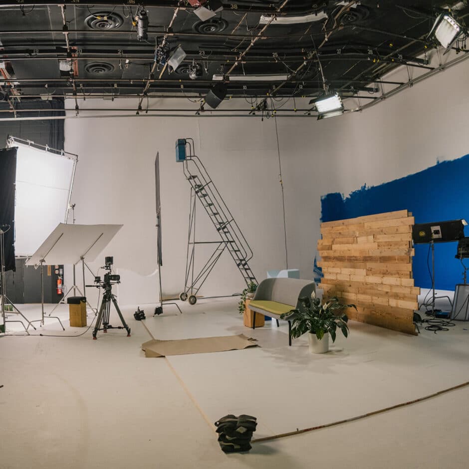 Film studio setup with overhead lights, tripods, and a large ladder. A wooden panel against a blue backdrop adds a rustic feel. Plants and a bench create a cozy vibe.