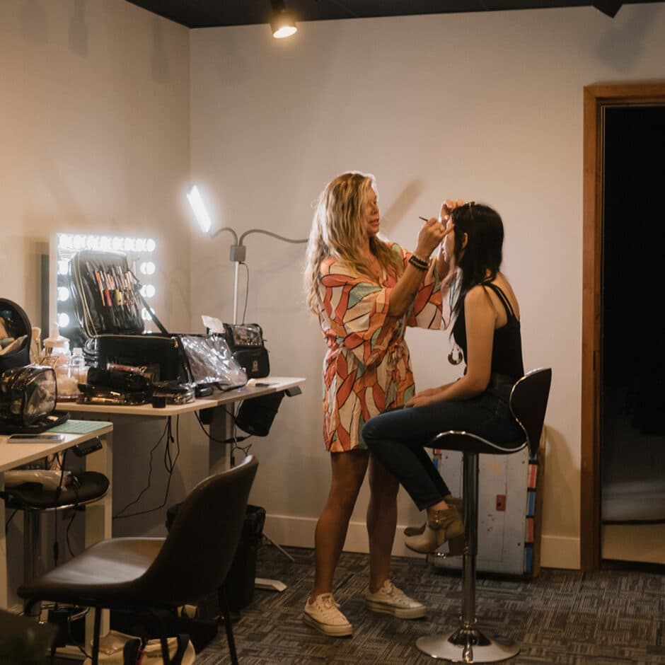 A makeup artist in a colorful dress applies makeup to a seated woman in a modern studio, surrounded by beauty tools and bright lighting, conveying focus and creativity.