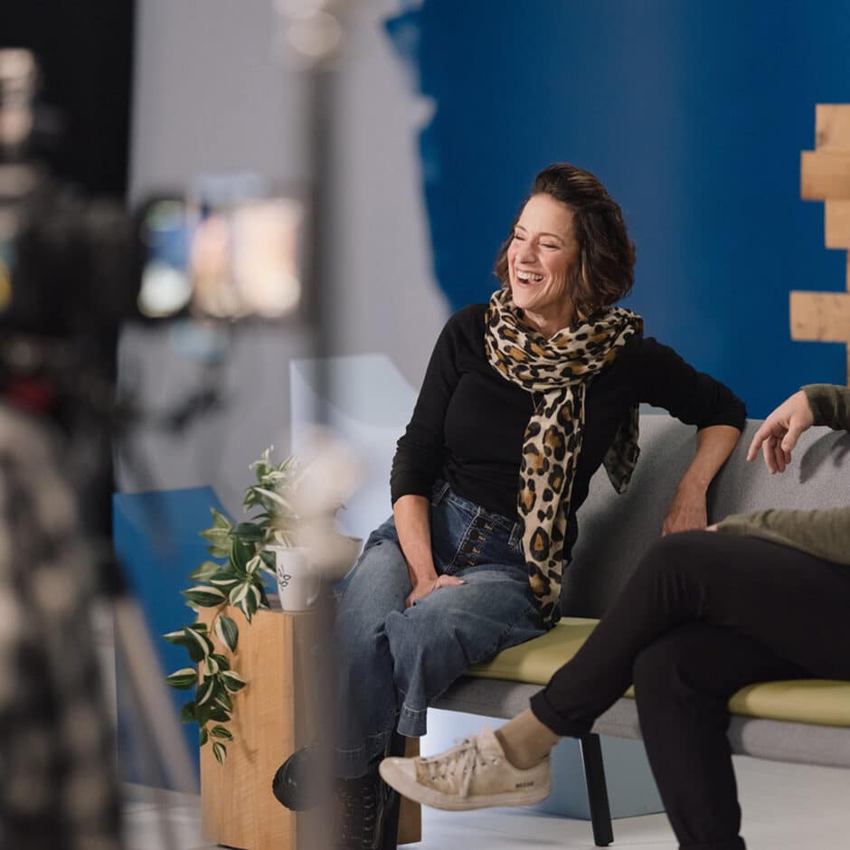 A woman in a black shirt and leopard print scarf laughs while sitting on a couch in a casual studio setting. A plant and camera are in the foreground.