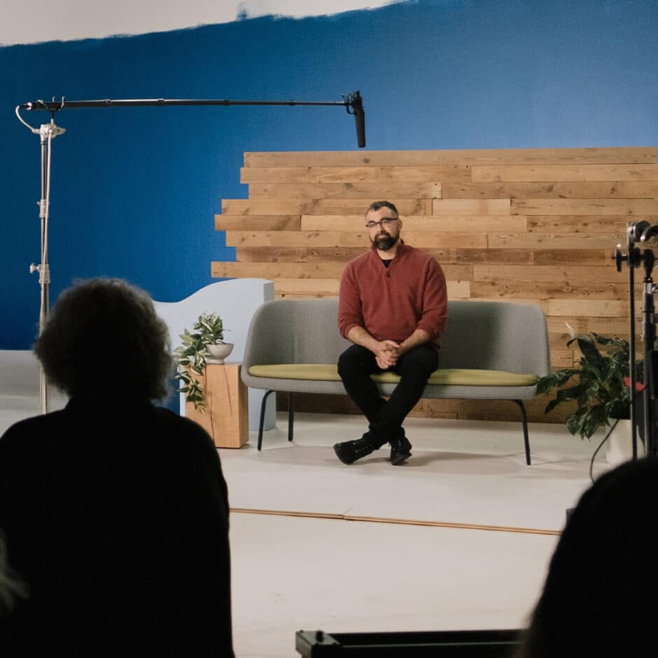A man in a red sweater sits on a gray couch in a studio with wooden paneling and a blue wall. A microphone hangs above, and plants decorate the scene.