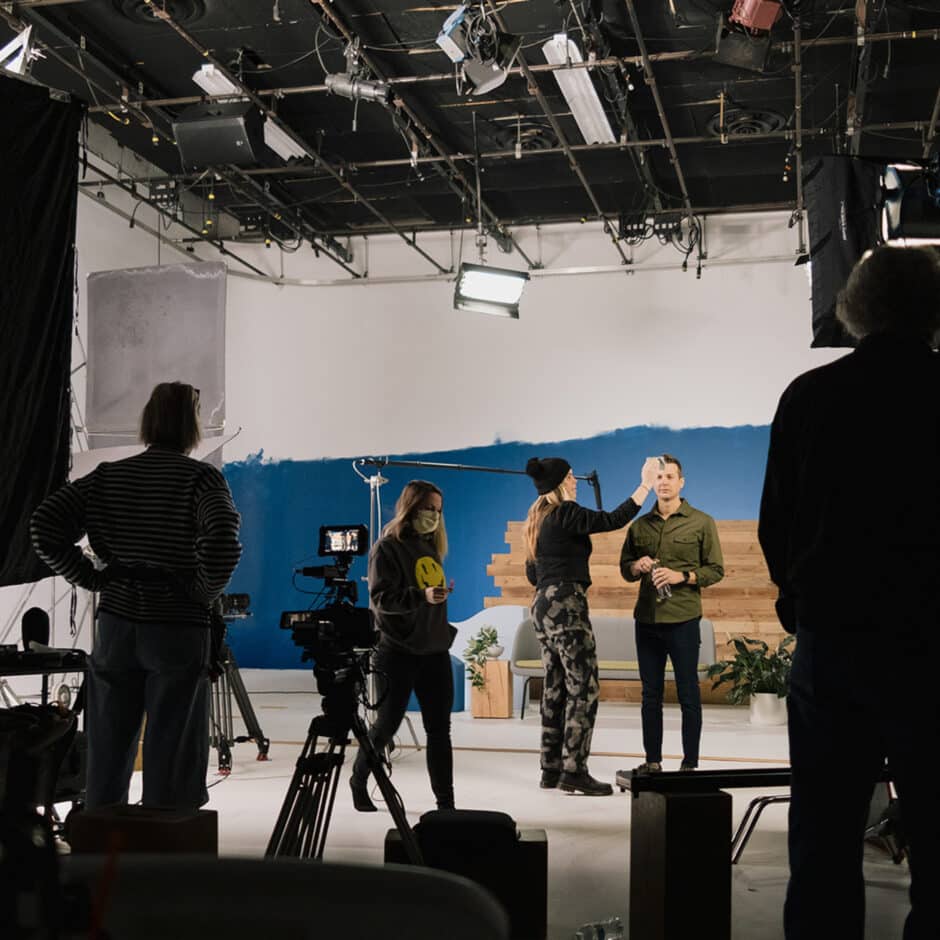 A film crew works in a studio with blue and white walls. A person adjusts the hair of a presenter under bright lights, while cameras and crew surround them.
