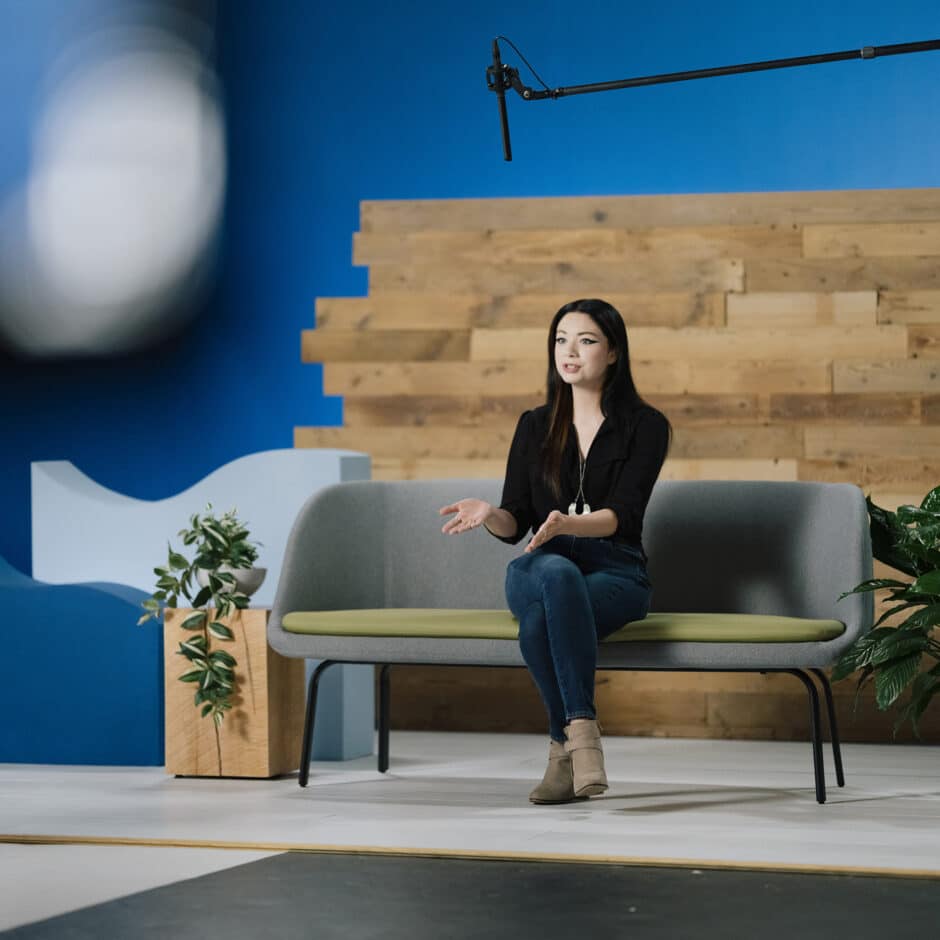 A woman sits on a modern grey couch in a studio setting with a wooden backdrop and blue wall. She's mid-conversation, with a microphone overhead.