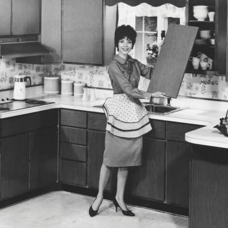 Black and white image of a smiling woman in a 1950s kitchen. She wears a polka-dot apron, holding an open cabinet door. Retro decor exudes a cheerful tone.