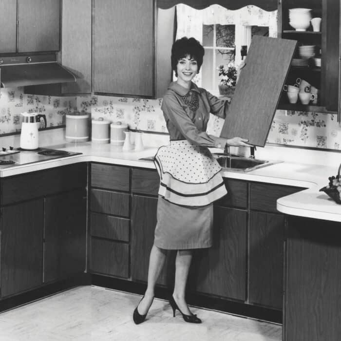Black and white image of a smiling woman in a 1950s kitchen. She wears a polka-dot apron, holding an open cabinet door. Retro decor exudes a cheerful tone.