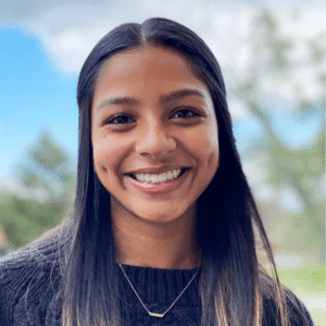 Smiling woman with long hair, wearing a black sweater and necklace. She stands outside with a blurred background of trees and blue sky, conveying a cheerful, relaxed mood.