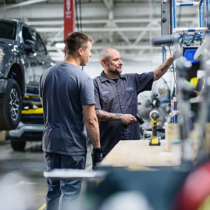 Two men stand in an auto repair shop. One points at equipment on a workbench, while a car is lifted in the background. The atmosphere is focused and collaborative.
