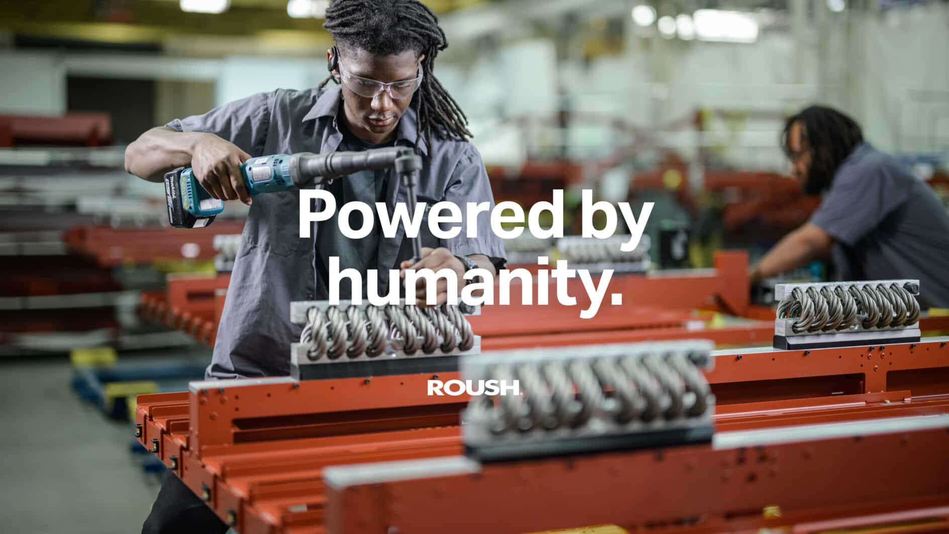 A focused worker uses a power tool on machinery in an industrial setting. Text overlaid reads "Powered by humanity." The atmosphere conveys innovation and teamwork.