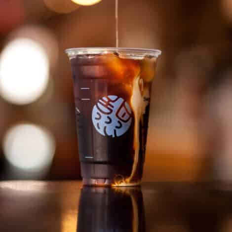 Iced coffee in a clear plastic cup with cream swirling in, sitting on a dark surface. Background is softly blurred with warm lights. Relaxing and inviting tone.