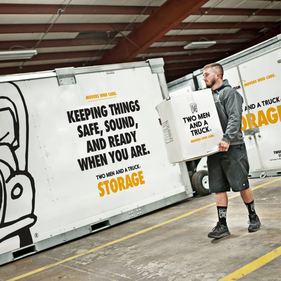 A man in casual attire carries a box labeled "Two Men and a Truck" inside a warehouse. Nearby, a large white storage container displays the slogan, "Keeping things safe, sound, and ready when you are." The atmosphere suggests professionalism and reliability.